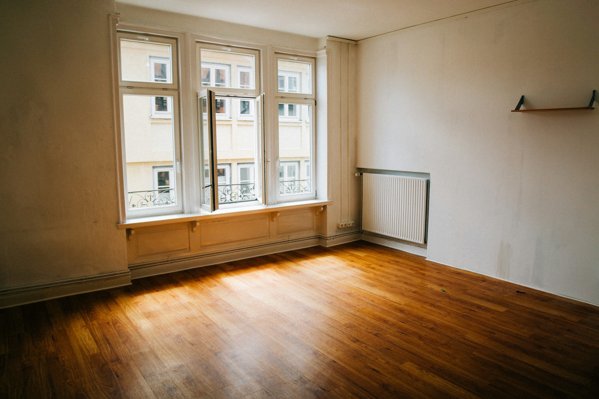 An empty Altbau room after renovation: tall double windows onto a Gründerzeit facade with wrought-iron balconies, panelled window niche and warm wooden floorboards in the afternoon sun