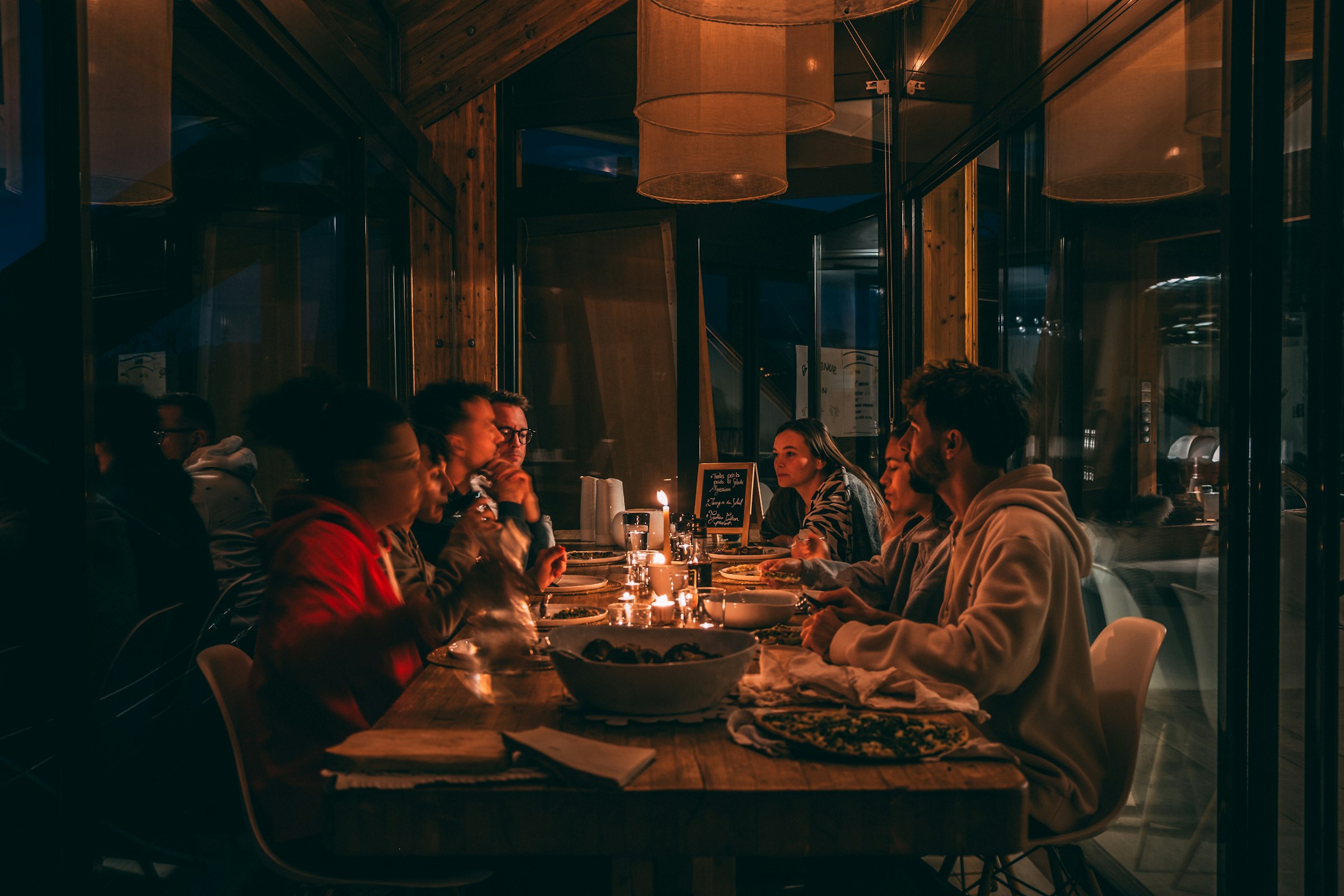 A group of friends eating at a long wooden table by candlelight, under fabric lampshades, with flatbread and a big bowl in the middle