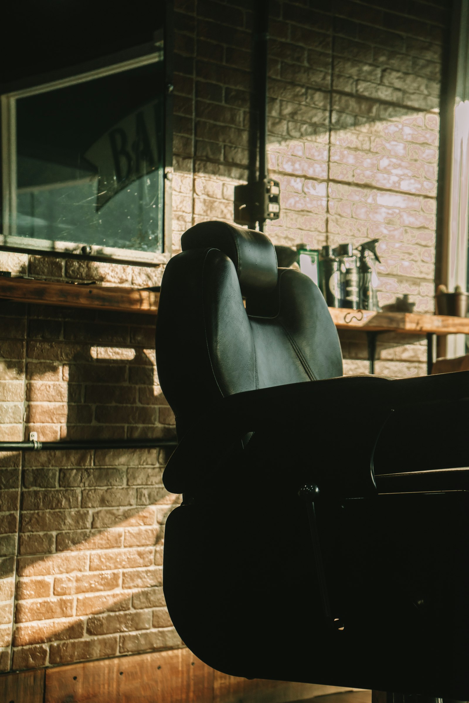 An empty black barber chair in low afternoon sun, against an exposed brick wall with a wooden shelf of tonics and a mirror